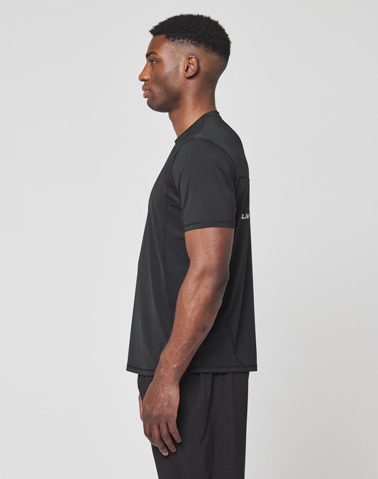 A man stands in profile facing left, wearing the LIVE FAST DIE YOUNG Live Fast Performance Tee in black and black pants against a plain white background.