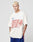 A person with short, bleached blonde hair wears the LFDY Juveniles Tee by LIVE FAST DIE YOUNG, featuring red and blue graphics, paired with blue athletic shorts with red and white stripes, against a plain light background.