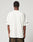 A person with short curly hair is seen from behind in a loose white LFDY Angel Tee by LIVE FAST DIE YOUNG (100% Baumwolle, black front and sleeve print) and dark pants, against a plain light background.