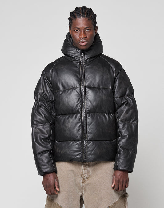 A person stands against a plain background wearing the LIVE FAST DIE YOUNG Live Fast Leather Puffer Jacket—black, oversized with a hood and embossed logo—paired with light brown, loose-fitting pants and neatly braided hair.