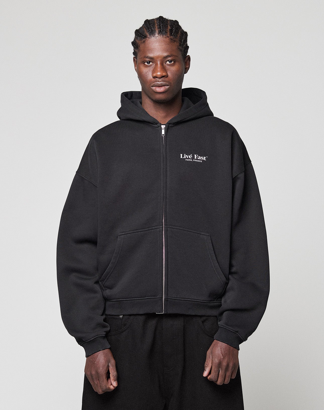 A person stands against a light gray background wearing the LIVE FAST DIE YOUNG LFDY Paris Hooded Zip, a black oversized zip-up hoodie made from Baumwolle Polyester with "Live Fast" in white on the left chest and a front pocket.