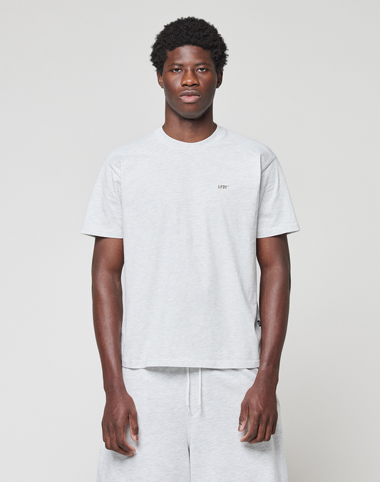 A man stands against a plain light background, wearing a light gray Basic Chest Tee by LIVE FAST DIE YOUNG and matching gray shorts. This regular-fit T-shirt is made from 100% Baumwolle and features small text on the left chest.