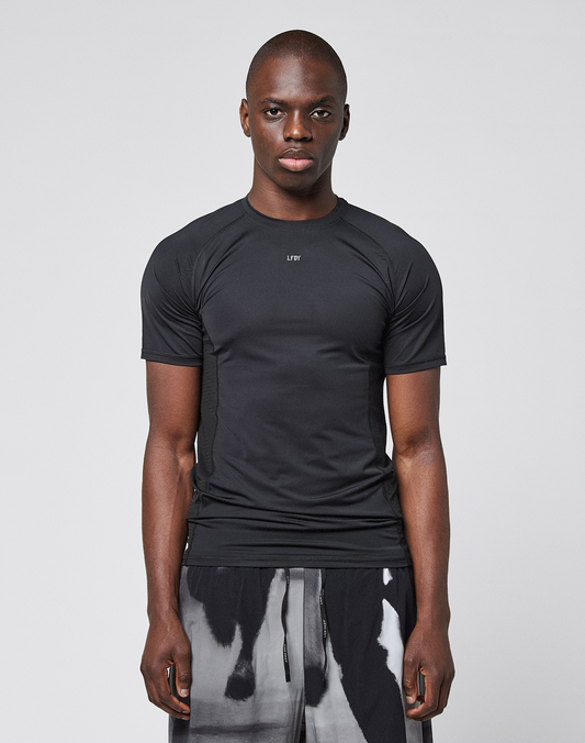 A person stands facing forward against a plain light gray background, wearing the LFDY Performance Slim Tee by LIVE FAST DIE YOUNG with a reflective "LIFY" logo print, paired with black-and-white patterned shorts.