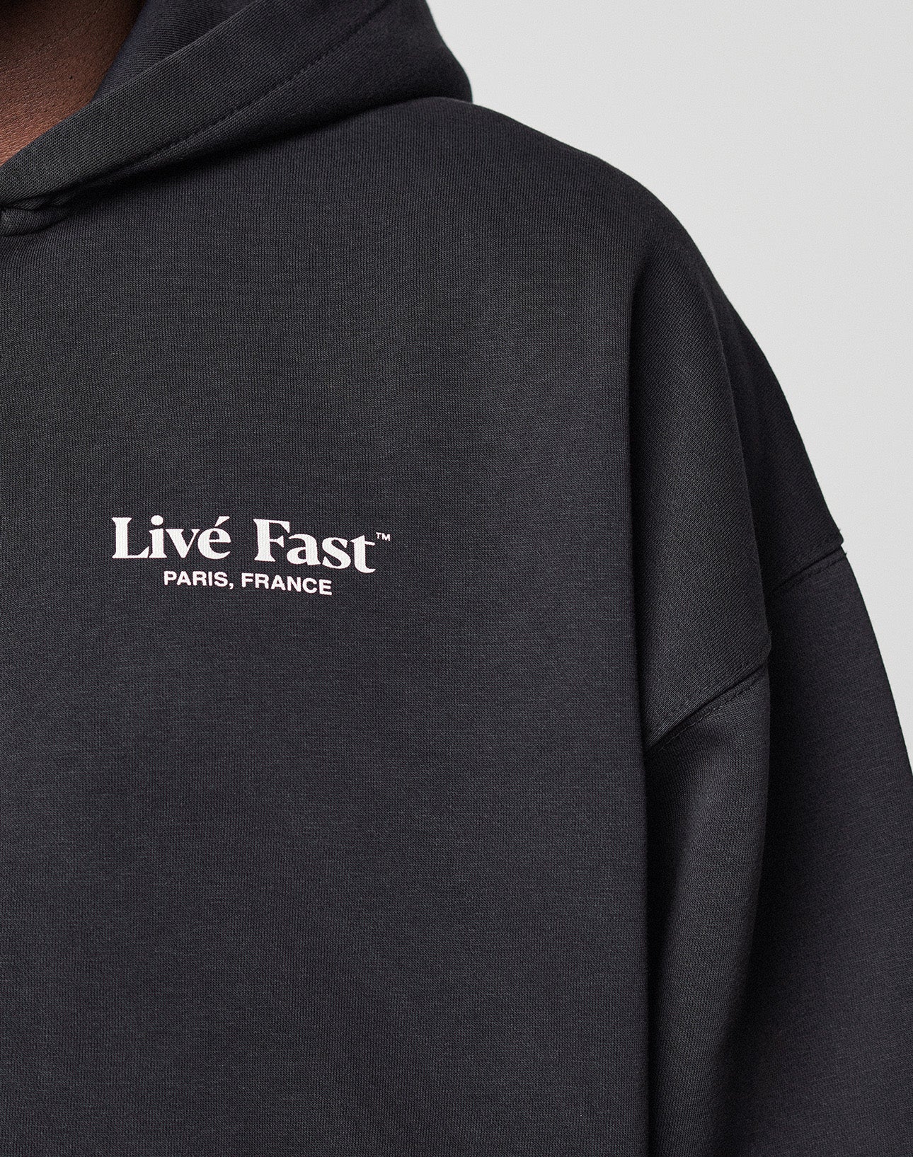 A close-up of the black LFDY Paris Hooded Zip by LIVE FAST DIE YOUNG, featuring front and back "Live Fast PARIS, FRANCE" prints in white on the left chest. The oversized cotton-polyester blend adds comfort; wearer stands before a light gray background.