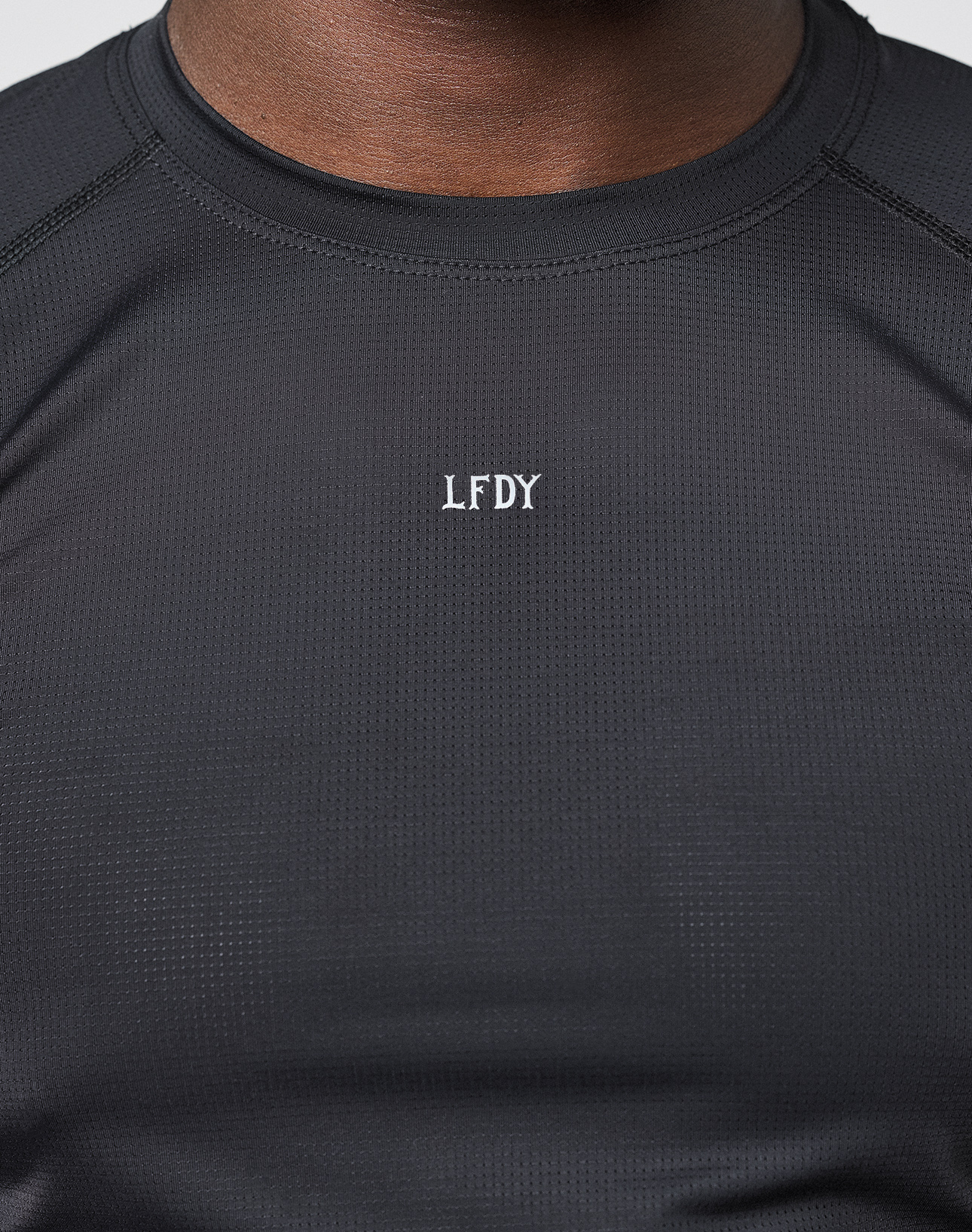 A close-up of someone wearing the LIVE FAST DIE YOUNG LFDY Performance Slim Tee in black, featuring white “LFDY” embroidery on the chest. The wearer’s face is not visible and the background is neutral.