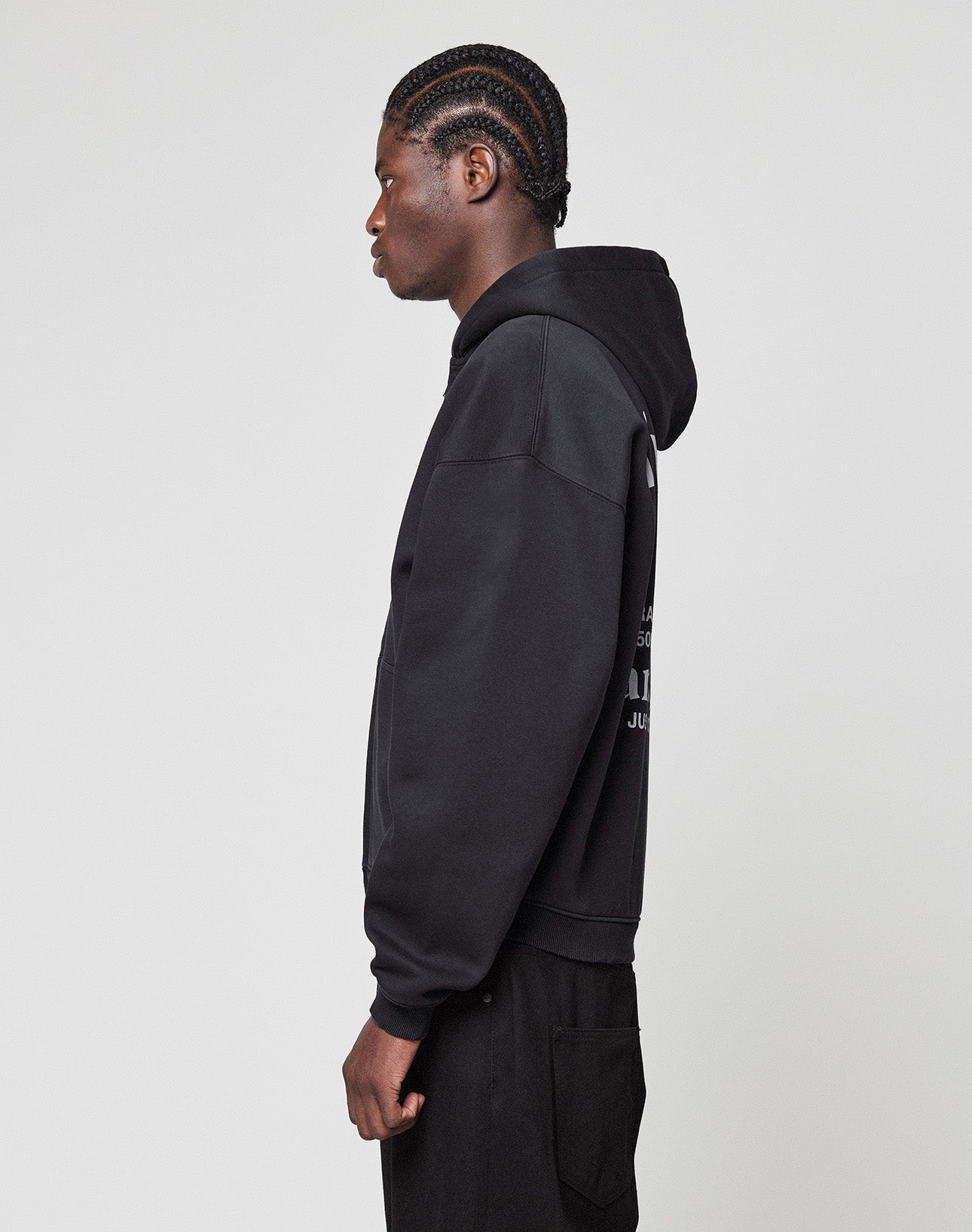 A person with cornrow braids stands in profile, wearing an oversized black LIVE FAST DIE YOUNG LFDY Paris Hooded Zip made from Baumwolle Polyester and black pants, set against a plain light gray background.