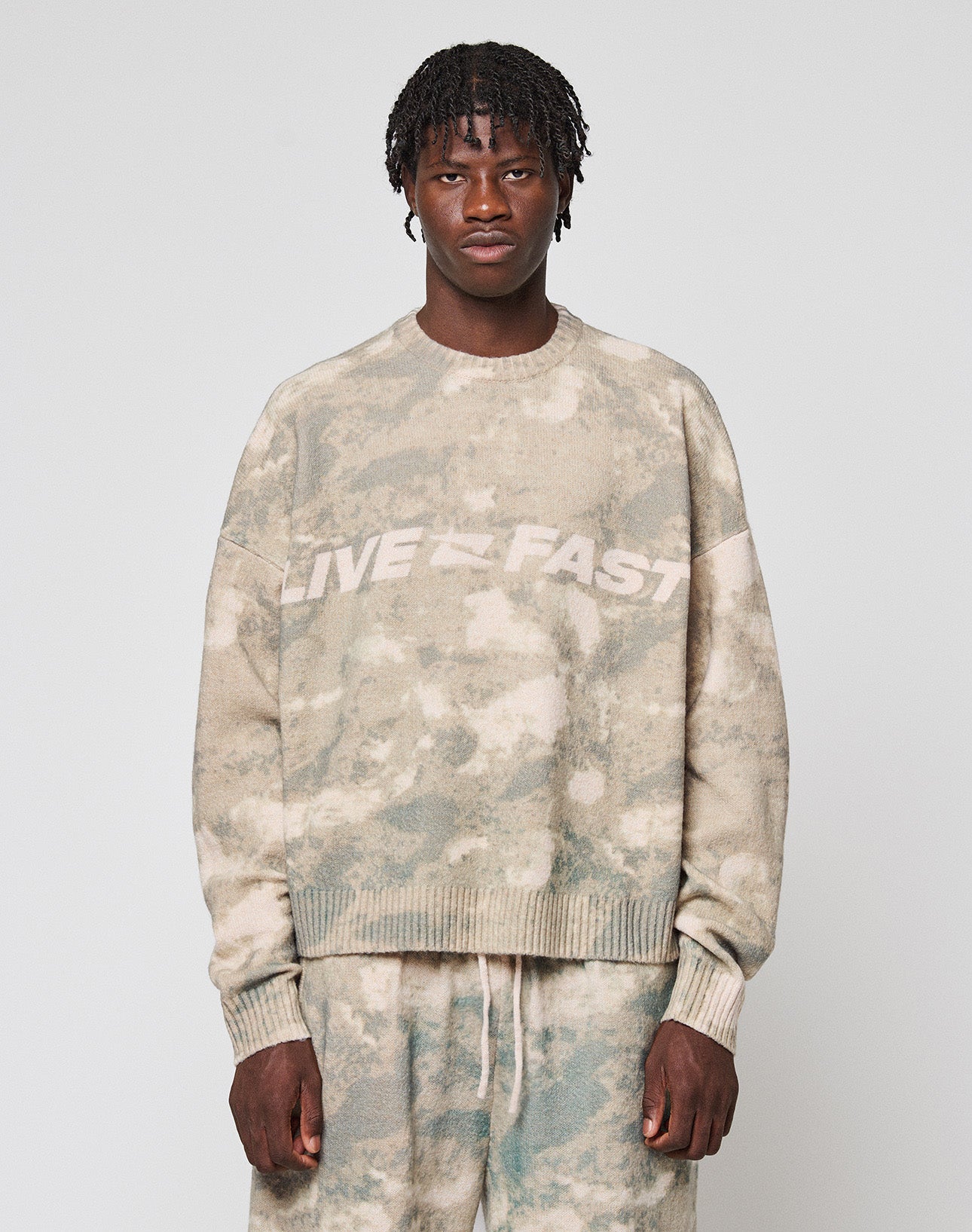 A person faces forward wearing the LIVE FAST DIE YOUNG Live Fast Boucle Sweater in an oversized beige and green tie-dye design with "LIVE FAST" on the chest, paired with matching soft polyester pants against a plain light gray background.