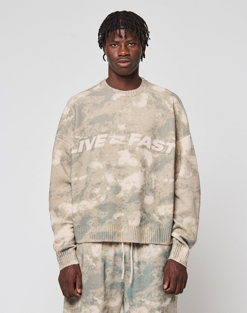 A person faces forward wearing the LIVE FAST DIE YOUNG Live Fast Boucle Sweater in an oversized beige and green tie-dye design with "LIVE FAST" on the chest, paired with matching soft polyester pants against a plain light gray background.