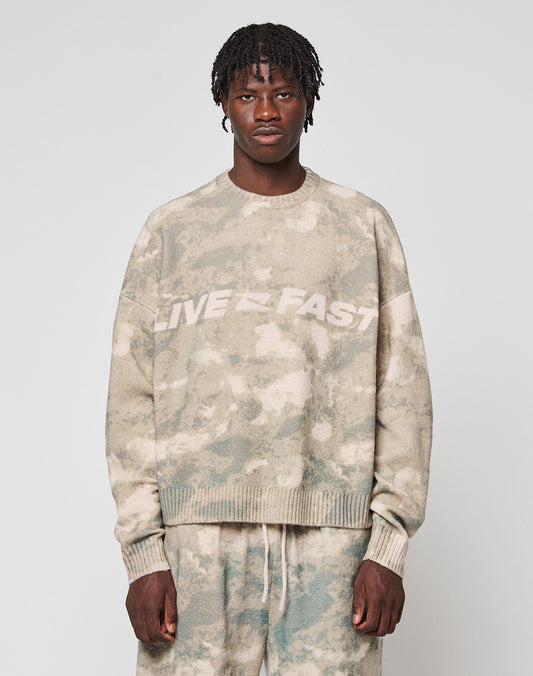 A person faces forward wearing the LIVE FAST DIE YOUNG Live Fast Boucle Sweater in an oversized beige and green tie-dye design with "LIVE FAST" on the chest, paired with matching soft polyester pants against a plain light gray background.