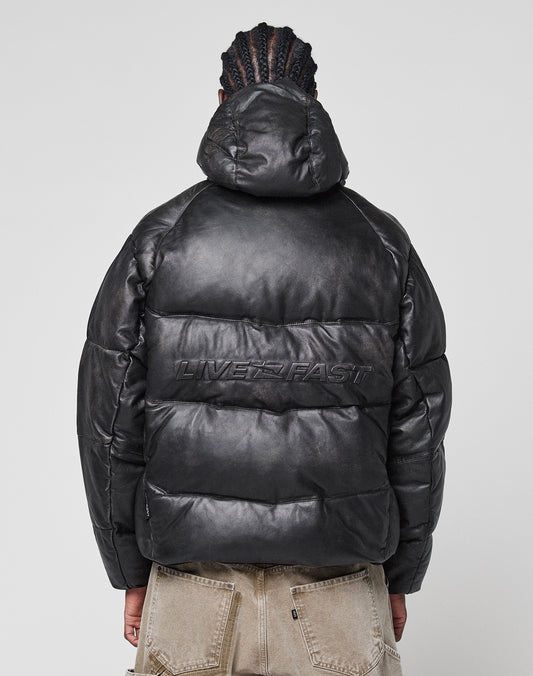 A person with braided hair, seen from behind, wears the LIVE FAST DIE YOUNG Live Fast Leather Puffer Jacket in black with an embossed logo on the back, a hood, and beige pants.
