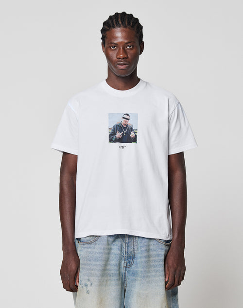 A man with braided hair stands against a plain background, wearing the LIVE FAST DIE YOUNG LFDY Gambling Tee—white, 100% Baumwolle, featuring a frontprint photo and text—paired with light blue loose-fitting jeans.