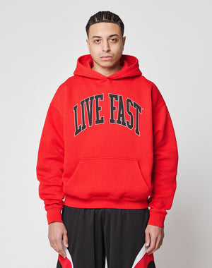A person faces the camera in an oversized bright red Live Fast College Hooded by LIVE FAST DIE YOUNG, featuring bold black LIVE FAST text and black/red athletic shorts, made from a comfortable polyester blend, against a plain white background.