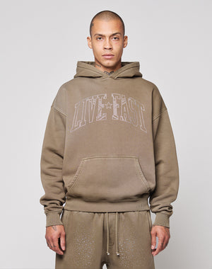 A person wears the LIVE FAST DIE YOUNG Live Fast Rhinestone Hooded, an oversize brown hoodie with bold “LIVE FAST” lettering on the front, standing against a plain light background with a neutral expression and hands by their sides.