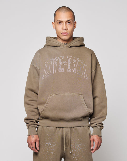 A person wears the LIVE FAST DIE YOUNG Live Fast Rhinestone Hooded, an oversize brown hoodie with bold “LIVE FAST” lettering on the front, standing against a plain light background with a neutral expression and hands by their sides.