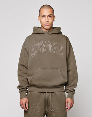 A person faces the camera against a plain background, wearing the LIVE FAST DIE YOUNG Live Fast Rhinestone Hooded in olive green with an oversize fit and "LIVE FAST" on the front, paired with matching speckled sweatpants.