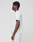 A person with short curly hair stands in profile facing left, wearing a light gray LIVE FAST DIE YOUNG Basic Chest Tee and matching shorts, both made from 100% Baumwolle, against a plain light background.