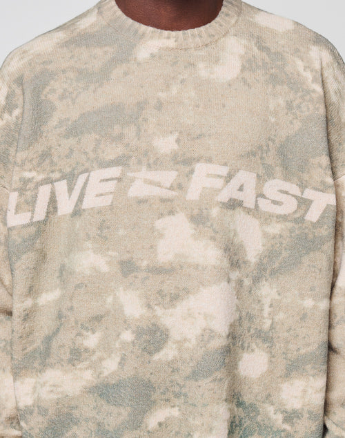 A person wears the Live Fast Boucle Sweater by LIVE FAST DIE YOUNG, featuring an oversized fit in beige and light green tie-dye with "LIVE FAST" in bold white letters across the chest. Only their lower face and upper torso are visible.