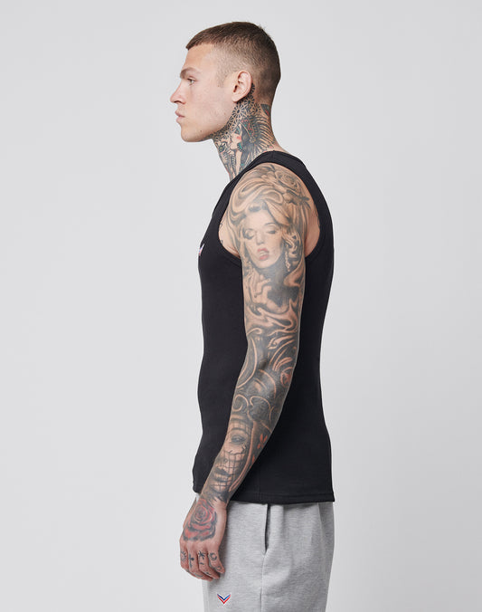 A tattooed man stands in profile facing left, wearing a black LFDY x Trigema Rib Tank by LIVE FAST DIE YOUNG and gray sweatpants against a plain light background.