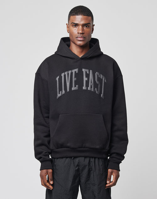 A man faces forward in a black LIVE FAST DIE YOUNG Live Fast College Hooded hoodie with large gray LIVE FAST text on the chest and black pants, standing against a plain light gray background.