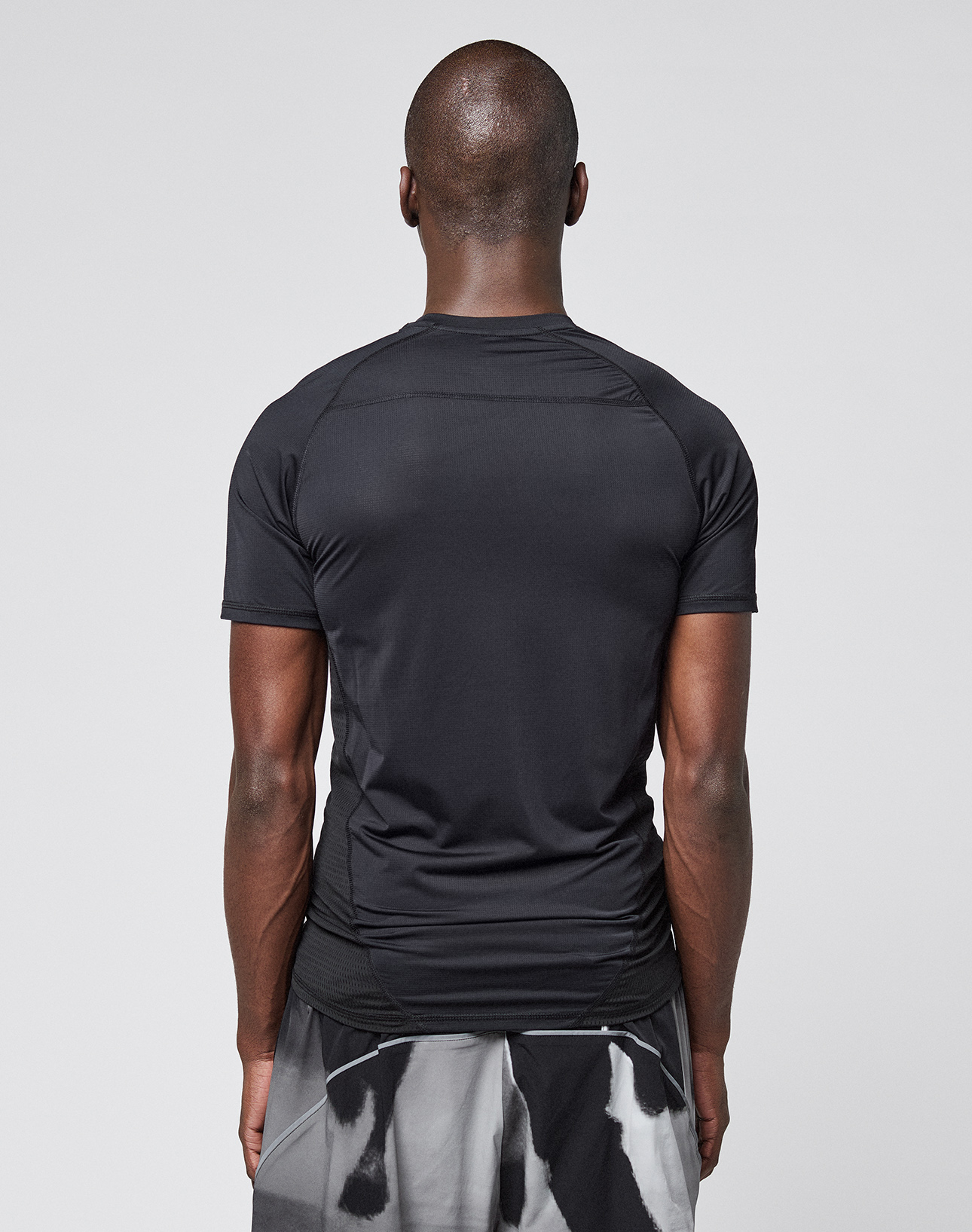 A person with short hair, seen from behind, wears the LFDY Performance Slim Tee by LIVE FAST DIE YOUNG and black-and-white patterned shorts against a plain light gray background.