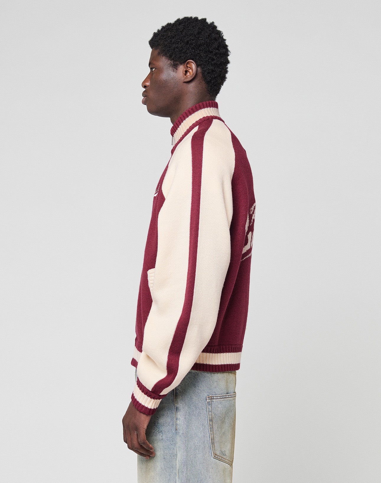 A person stands in profile wearing the LIVE FAST DIE YOUNG Boxy Knit Zip Jacket with an oversize fit and light blue jeans against a plain light gray background.