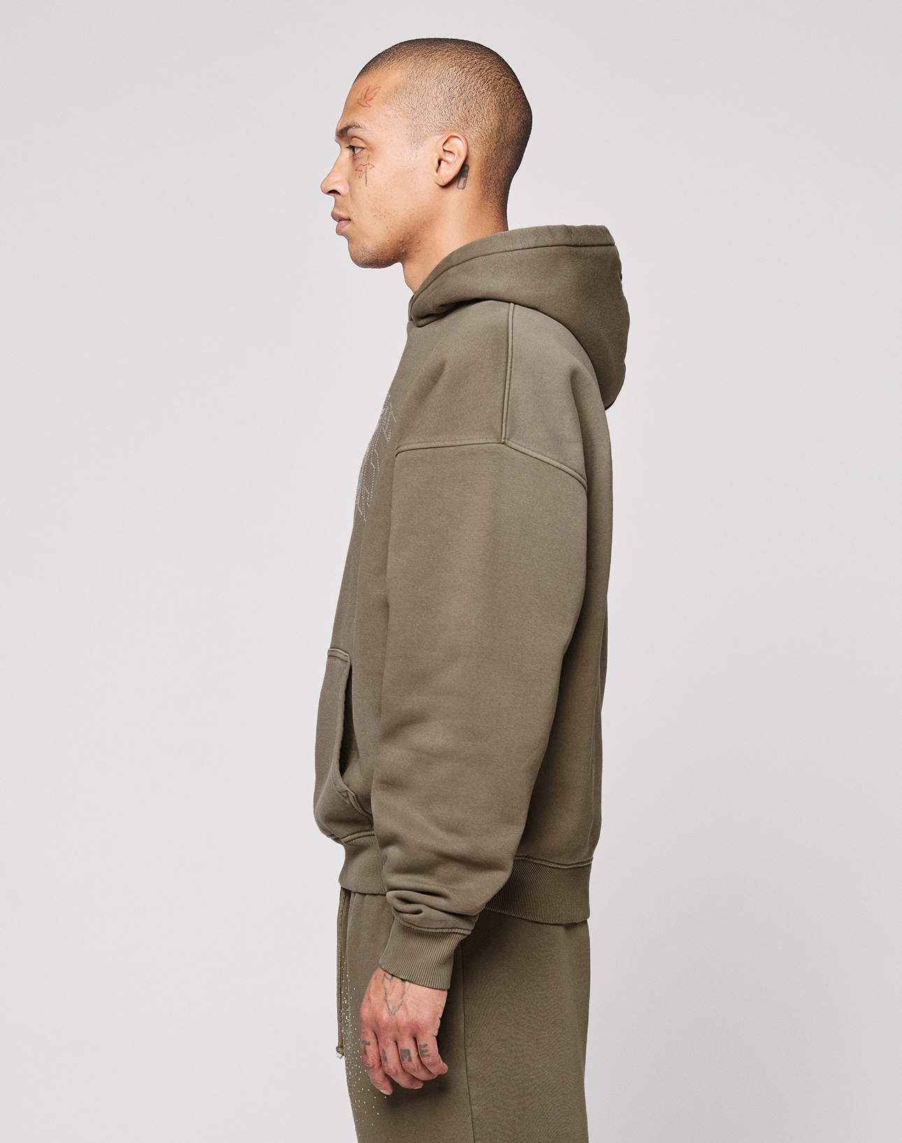 A person with close-cropped hair stands in profile, facing left, wearing the LIVE FAST DIE YOUNG Live Fast Rhinestone Hooded sweatshirt and matching oversized pants in olive green against a plain light gray background.