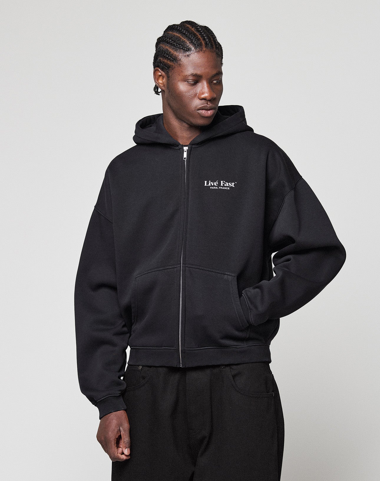 A person wears the LIVE FAST DIE YOUNG LFDY Paris Hooded Zip, a black oversized zip-up hoodie with "Live Fast" text on the chest, standing with one hand in their pocket against a plain light background.