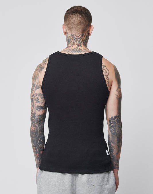 A person with tattooed arms and neck is shown from behind, wearing a LIVE FAST DIE YOUNG LFDY x Trigema Rib Tank in black and light gray shorts, standing against a plain light background.