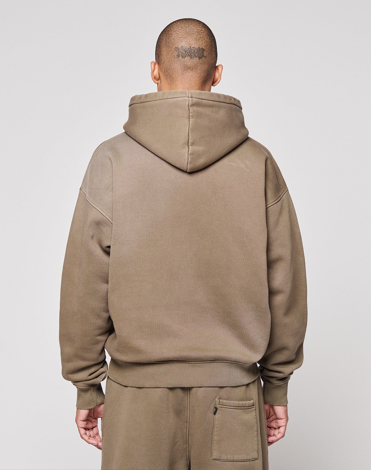 A person in the LIVE FAST DIE YOUNG Live Fast Rhinestone Hooded and matching sweatpants stands with their back to the camera, hood up, showing a tattoo on their shaved head. The plain background highlights the relaxed style.