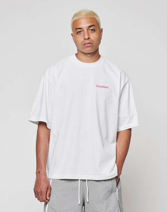 A person with short bleached blond hair wears the LIVE FAST DIE YOUNG Basic 280 Tee, a loose white 100% Baumwolle T-shirt with small pink “LiveFast” text on the chest, plus light gray drawstring shorts, standing against a plain light background.