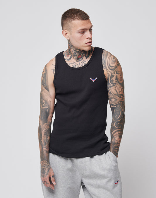 A tattooed young man wears the LIVE FAST DIE YOUNG LFDY x Trigema Rib Tank in black with gray sweatpants, standing with one hand in his pocket and looking to the side against a plain light background.