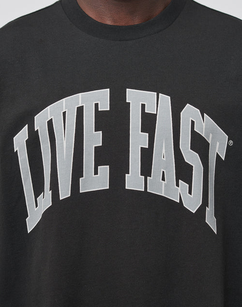 Someone wears the Live Fast College Tee by LIVE FAST DIE YOUNG, an oversized black shirt featuring a bold white LIVE FAST print across the chest.
