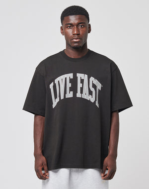 A man faces forward wearing the LIVE FAST DIE YOUNG Live Fast College Tee in oversize black 100% Baumwolle with bold white LIVE FAST text, paired with light gray shorts, against a plain light background.
