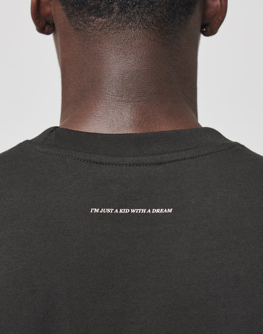 A person seen from behind wears the Live Fast College Tee by LIVE FAST DIE YOUNG—an oversize dark shirt featuring white “IM JUST A KID WITH A DREAM” text near the collar.