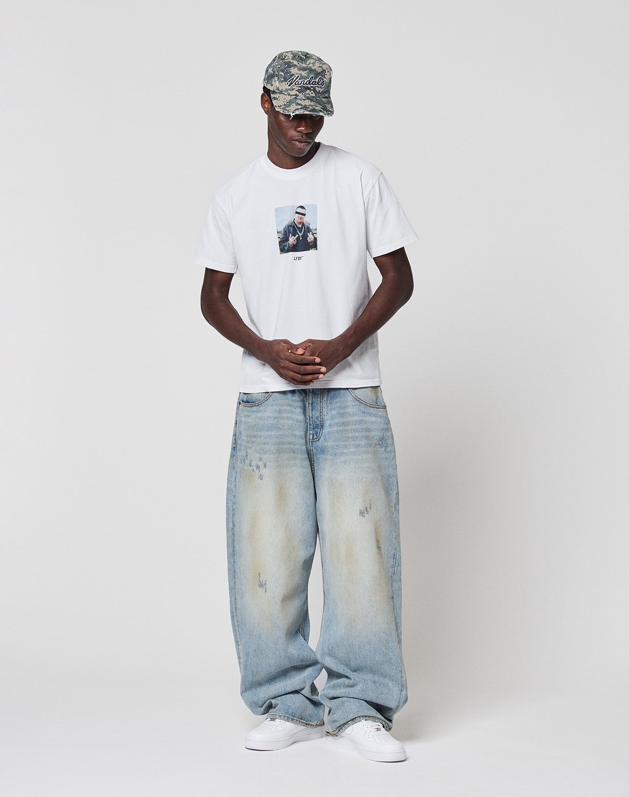 A man in a camouflage cap, light blue baggy jeans, white sneakers, and a LIVE FAST DIE YOUNG LFDY Gambling Tee stands against a plain white background with his hands clasped in front of him.