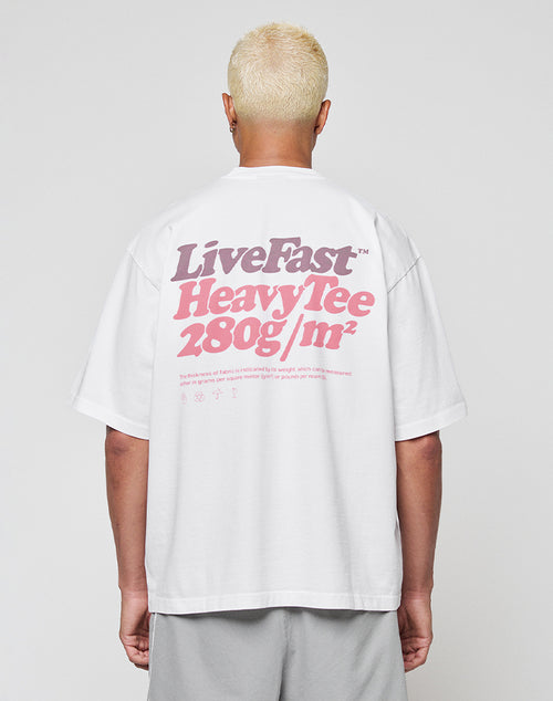 A person with short, bleached blonde hair faces away from the camera wearing the LIVE FAST DIE YOUNG Basic 280 Tee, an oversize white T-shirt made from 100% Baumwolle with a bold pink and gray backprint.