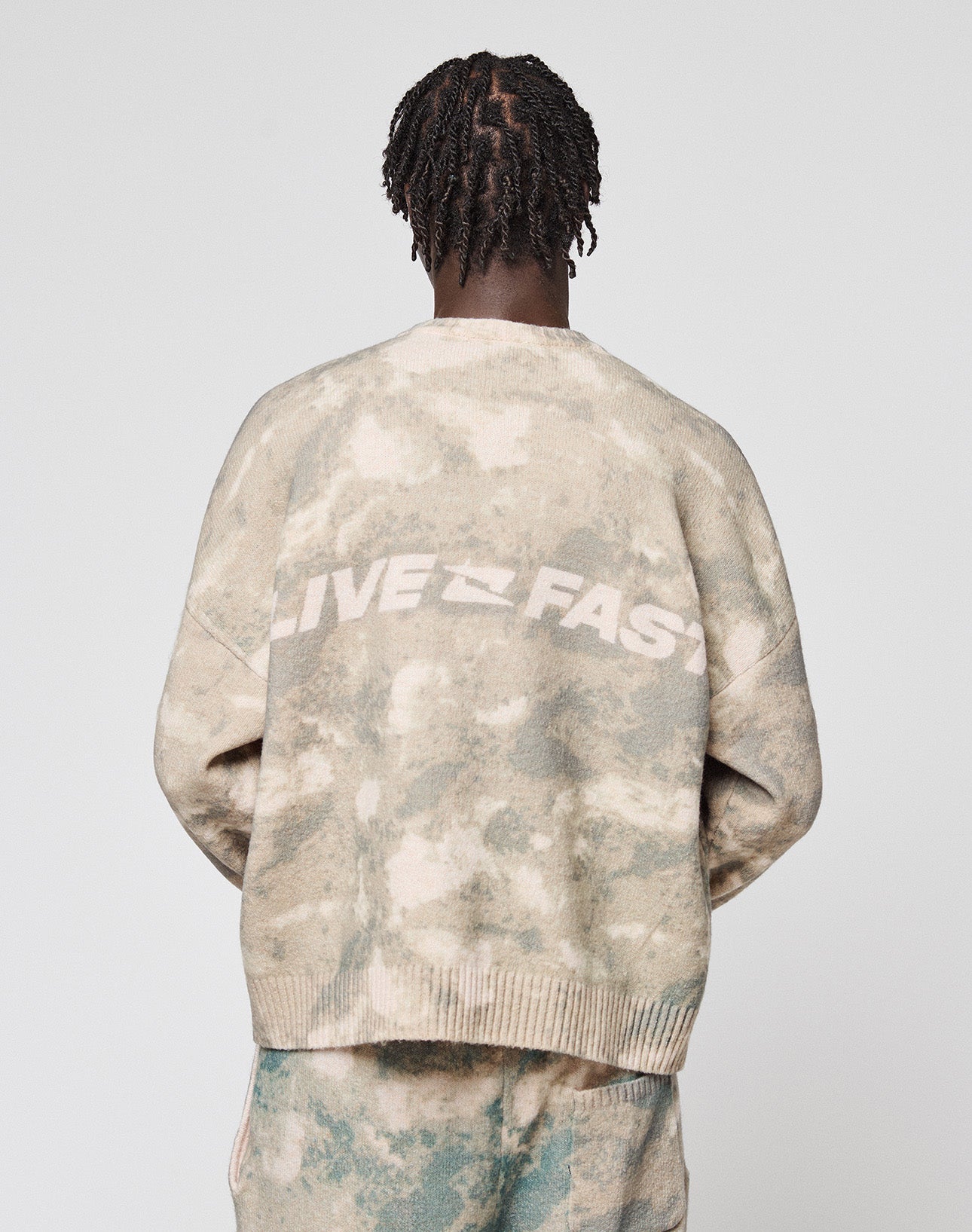 A person with short, twisted braids is seen from behind in an oversized beige and green tie-dye Live Fast Boucle Sweater by LIVE FAST DIE YOUNG, featuring "LIVE FAST" in white across the back against a plain light gray background.