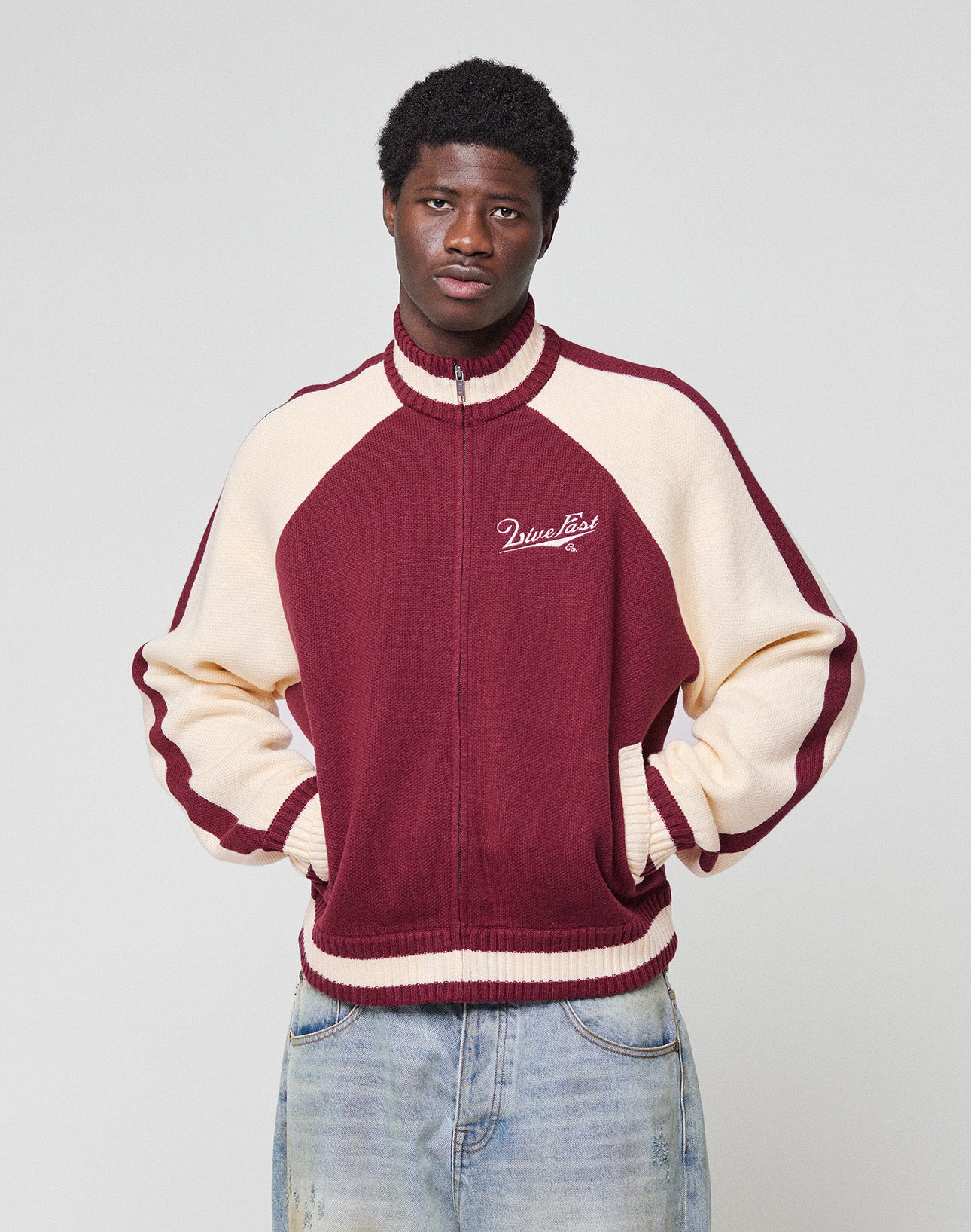 A man with short curly hair stands against a plain background, hands in pockets, wearing the LIVE FAST DIE YOUNG Boxy Knit Zip Jacket in maroon and cream with script on the chest, paired with light blue Oversize Fit jeans.