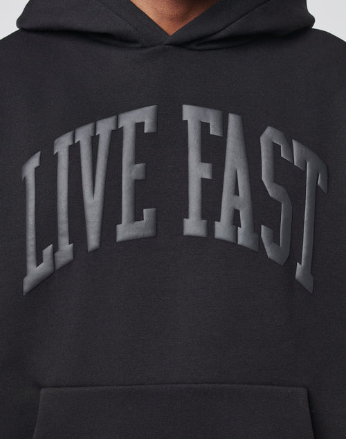 A close-up of the Live Fast College Hooded by LIVE FAST DIE YOUNG, a black hoodie with bold, raised dark gray LIVE FAST lettering across the chest. Designed with an oversize fit for a relaxed style.
