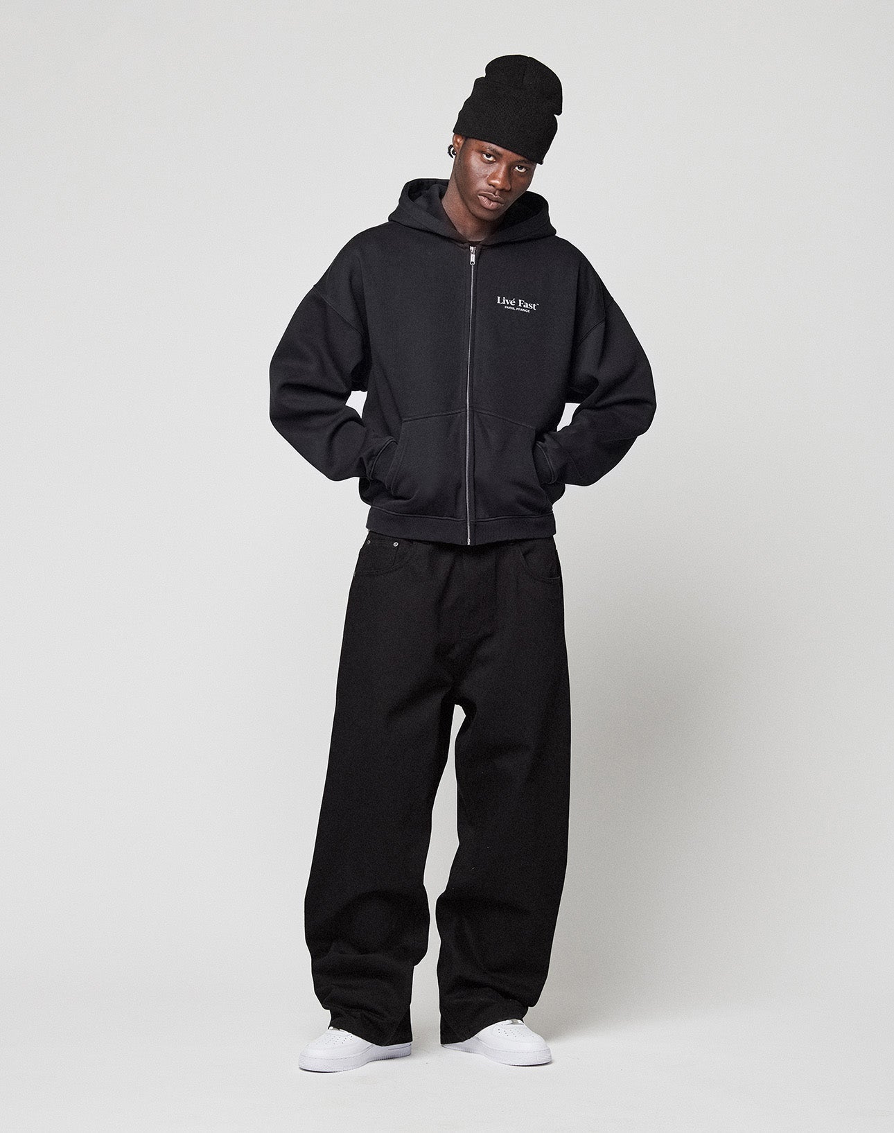 A person stands against a plain background wearing an oversized fit LIVE FAST DIE YOUNG LFDY Paris Hooded Zip in black, paired with a black beanie, baggy pants, and white sneakers, hands on hips and head slightly tilted.