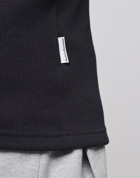Close-up of the side of a black LFDY x Trigema Rib Tank by LIVE FAST DIE YOUNG, featuring a small white Hergestellt in Deutschland tag, styled over light gray pants.