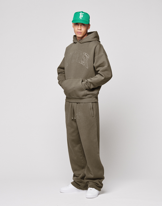 A person wears the LIVE FAST DIE YOUNG Live Fast Rhinestone Hooded in olive green with matching sweatpants, a green cap, and white sneakers, standing against a plain light background.