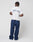 A person stands facing away, showcasing the LIVE FAST DIE YOUNG Live Fast Paris Tee, a regular fit white t-shirt with blue and red Live Fast front and back print, paired with loose navy pants and white shoes against a plain white background.