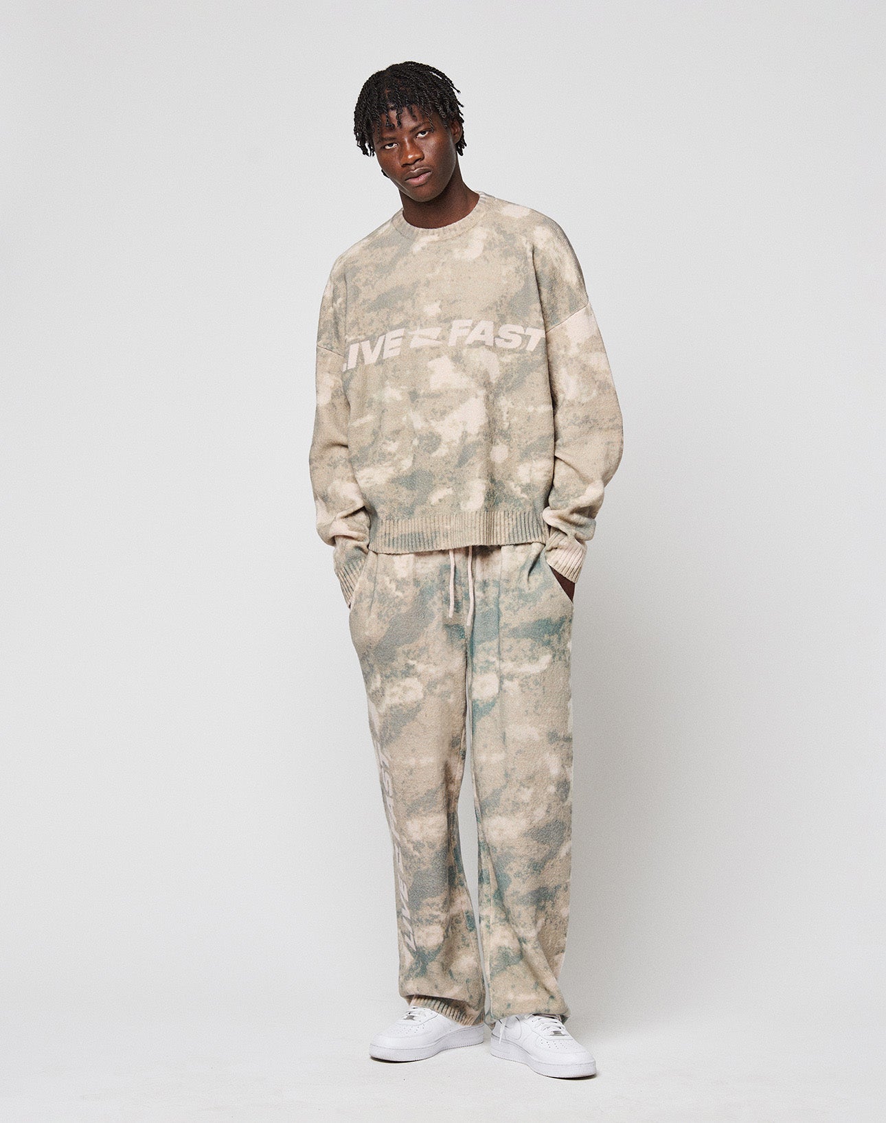 A person with short dreadlocks and a relaxed expression wears LIVE FAST DIE YOUNG’s Live Fast Boucle Pants, featuring a breiter Elastikbund and gerades Bein, paired with a matching beige-green tie-dye top and white sneakers.