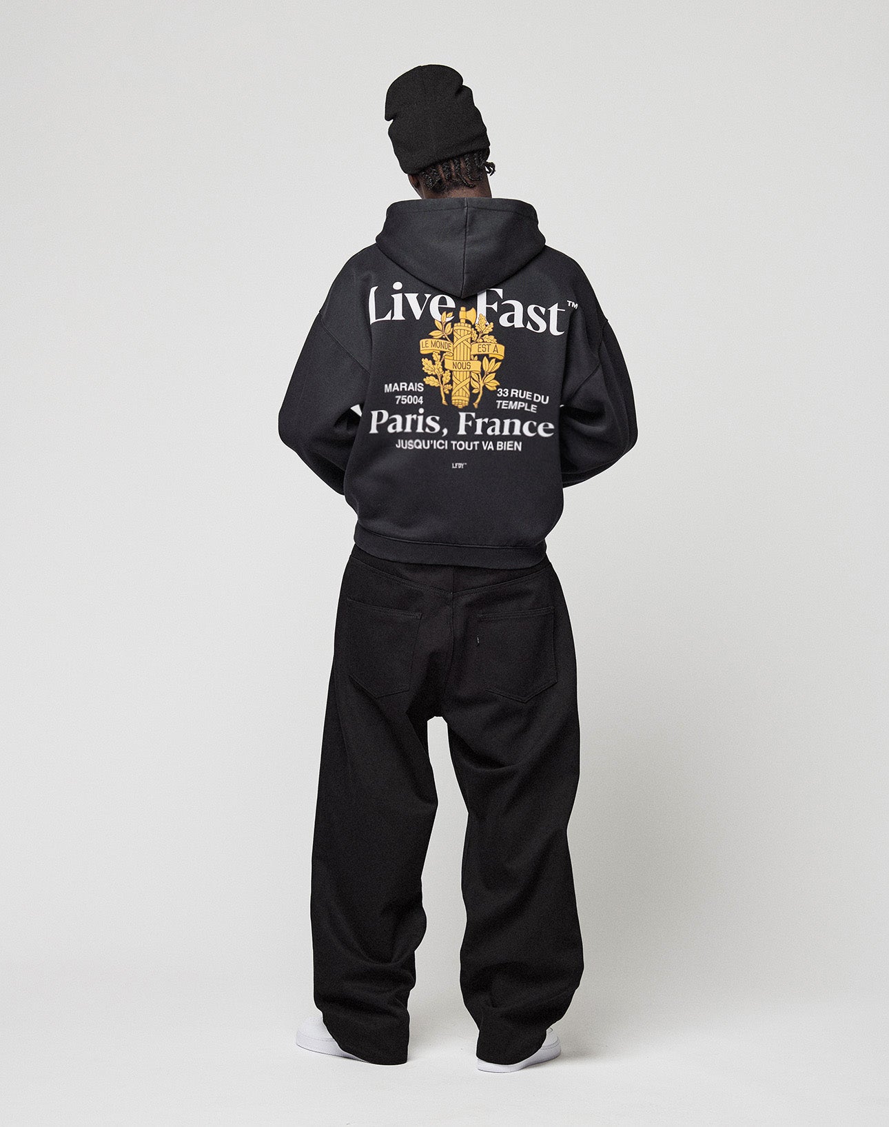 A person faces away wearing the LFDY Paris Hooded Zip by LIVE FAST DIE YOUNG, an oversized black Baumwolle-Polyester hoodie with Live Fast and Paris, France front and back prints, styled with black baggy pants, a black beanie, and white shoes.
