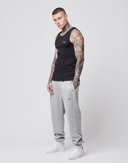 A tattooed man with short hair wears the LFDY x Trigema Rib Tank by LIVE FAST DIE YOUNG, paired with light gray sweatpants and white sneakers, standing against a plain white background and looking slightly to the side.