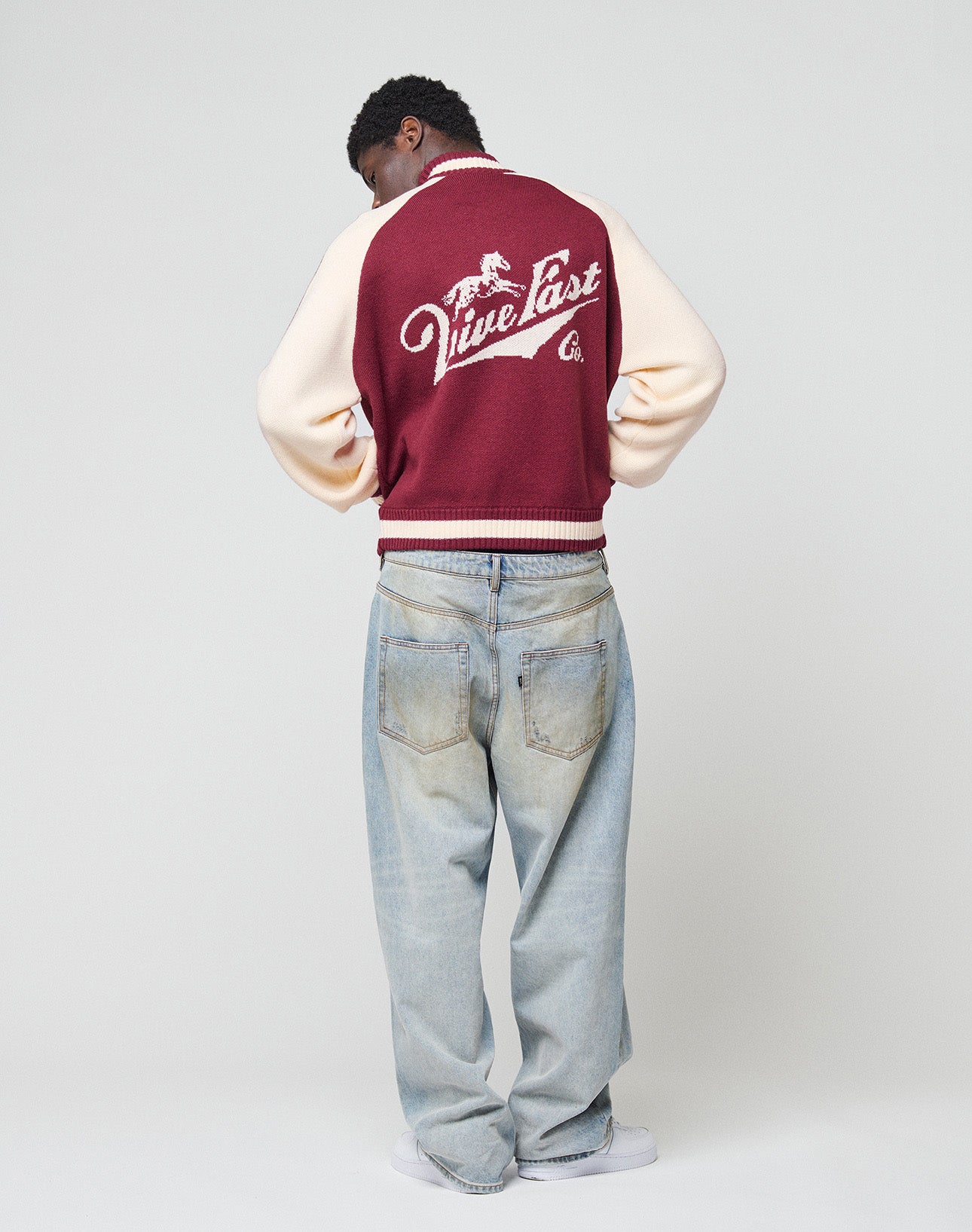 A person faces away, wearing a Boxy Knit Zip Jacket by LIVE FAST DIE YOUNG in burgundy and cream with a horse graphic and "Live Fast" on the back, styled with light blue baggy jeans and white sneakers against a plain background.