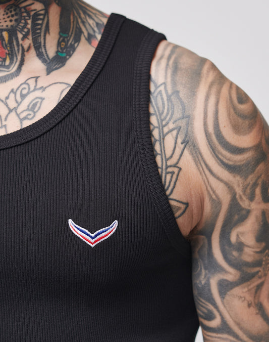 A close-up of a tattooed person wearing the LIVE FAST DIE YOUNG LFDY x Trigema Rib Tank, a slim fit black ribbed top with a small red, white, and blue embroidered logo on the chest.