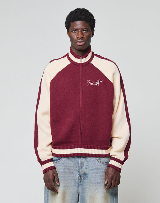 A young man with short curly hair and a neutral expression stands against a plain background, wearing the LIVE FAST DIE YOUNG Boxy Knit Zip Jacket in maroon and cream, featuring "MissList" on the chest, paired with light blue jeans.