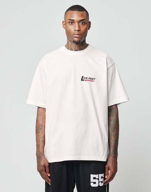 A tattooed man wears the LFDY Industries Tee by LIVE FAST DIE YOUNG, an oversized loose white cotton T-shirt with bold LIVE FAST prints front and back, plus black shorts with number 55, against a plain light background.